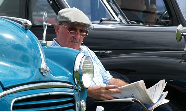 man in hat and glasses sitting between two classic cars and reading newspaper