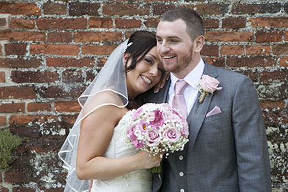 bride holding pink flowers with her head on the groom against an old brick wall background