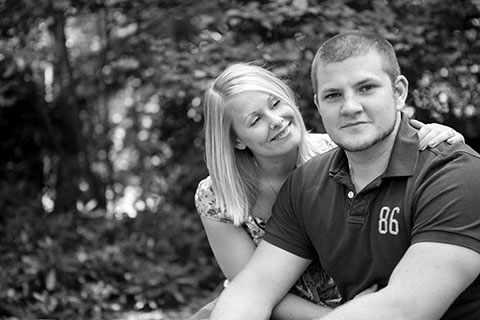 future bride with her hand on shoulder of groom and smiling as he looks straight at the camera in Essex woodlands