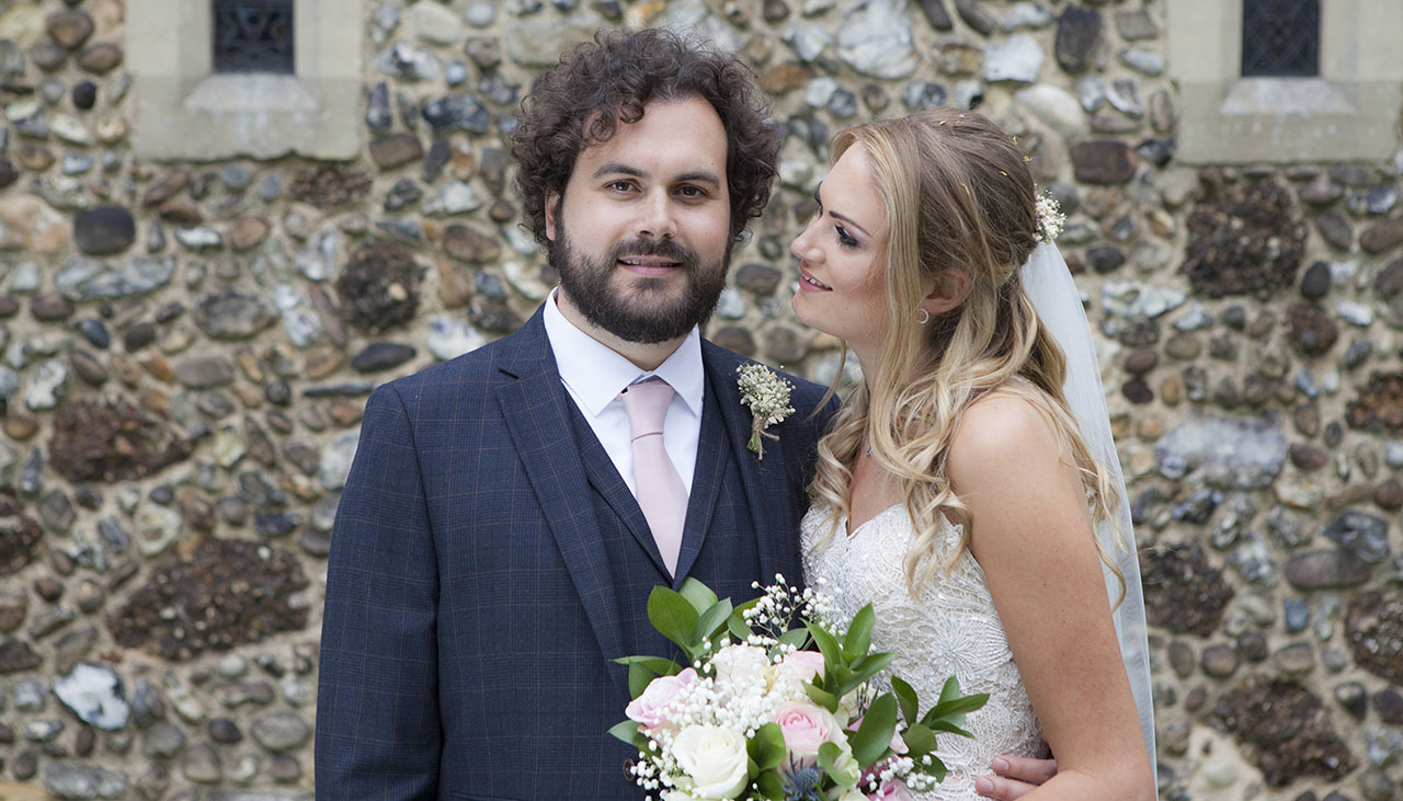 Groom looking at the camera standing next to his bride in front of a flint stone Essex church wall