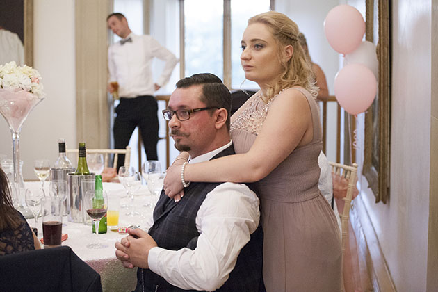 bridesmaid standing behind a seated wedding guest with her arms around his neck and shouders