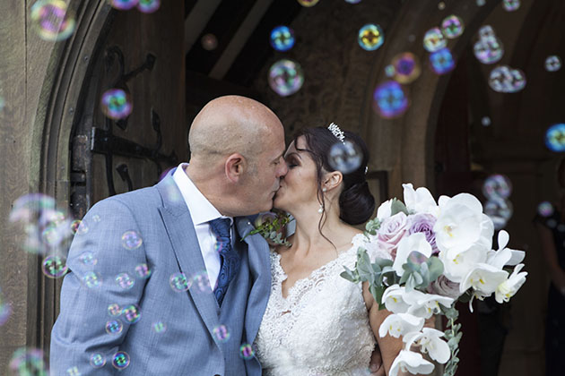 Bride and groom kissing in church doorway surrounded by bubbles